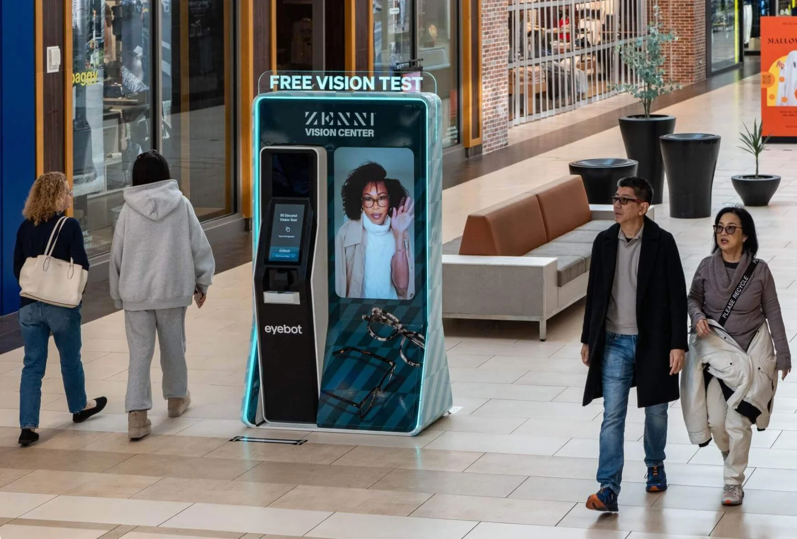 Shoppers walking past a Free Vision Test kiosk in a mall corridor.